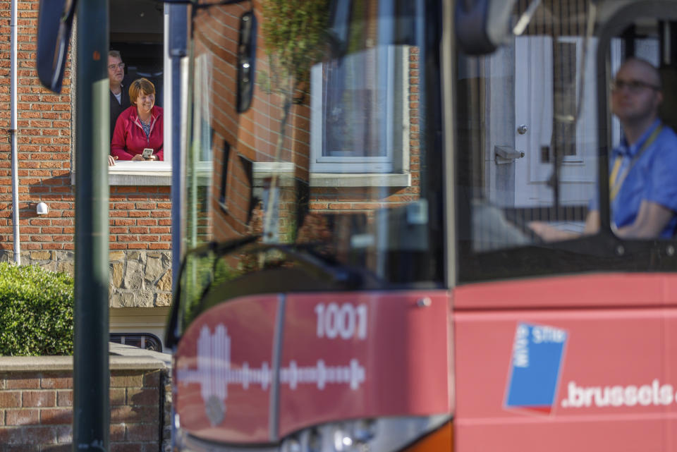 A couple lean out of their window to greet a bus delivering a loudspeaker message from family and friends in Brussels, Wednesday, April 22, 2020. With streets in the Brussels capital mostly devoid of loud traffic and honking horns, a simple piece of emotional poetry can split the air. The public bus company, STIB-MIVB, has been calling on people to send in voice messages, which are now delivered by a special bus driving in a loop to connect all the messages and leave a trail of happiness. (AP Photo/Olivier Matthys)