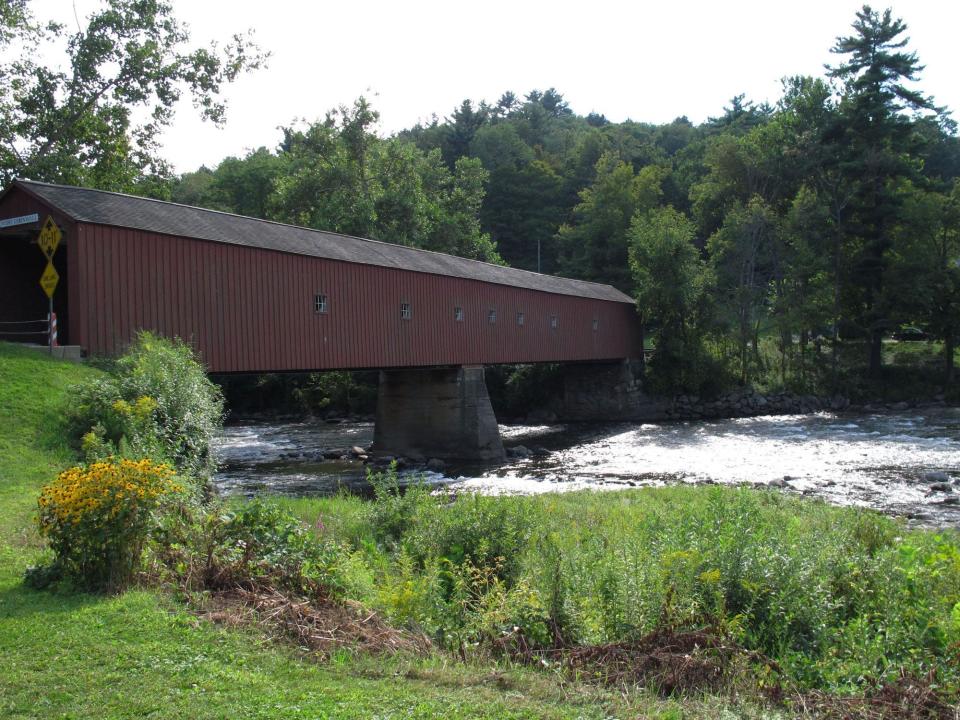 river runs under a red covered bridge with trees and greenery on either side