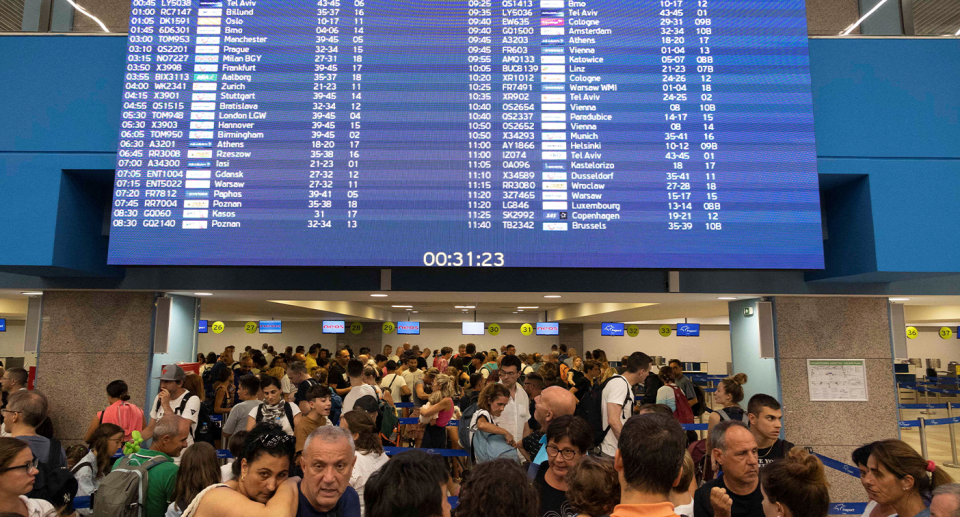 The airport in Rhodes. Dozens of people line up under the arrival and departure sign.