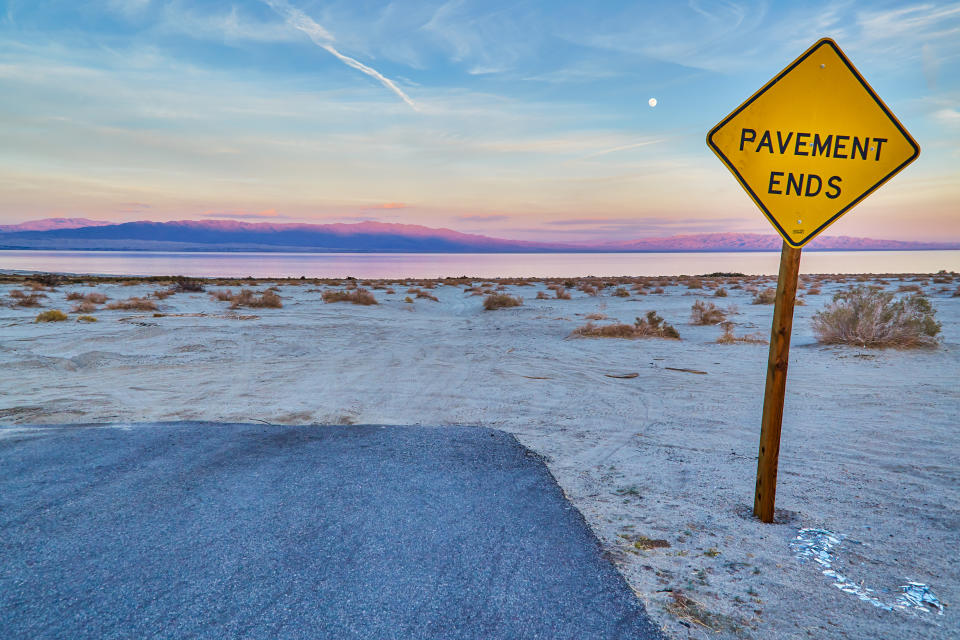 Sign reading "PAVEMENT ENDS" next to a road transitioning into a natural landscape at dusk