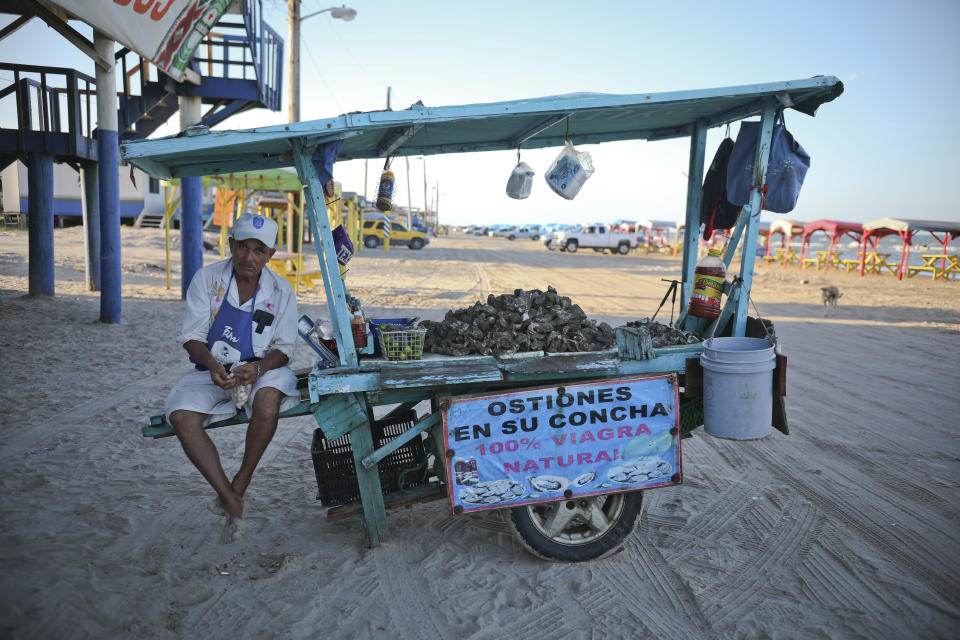 In this Aug. 2, 2019 photo, an oyster vendor snacks on shelled peanuts while he waits for customers in Playa Bagdad near the border city of Matamoros, Mexico. Here, the landscape is not one of walls or border guards; it is simply miles of dunes and Gulf coast beaches, marked only by simple wooden huts or awnings held up by sticks. (AP Photo/Emilio Espejel)