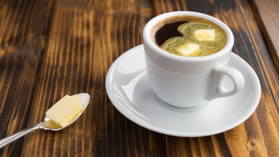 A cup of coffee with two spoons of butter melting into it, there's a spoon of butter on the side too