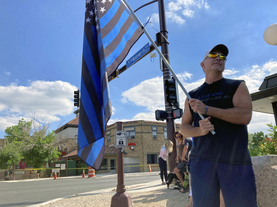 John Garrod, of Arvada, stands holding a blue line flag at the beginning of a line of about 30 police cars lined up for a procession in honor of the officer who was fatally shot in Arvada, Colo., on Monday, June 21, 2021. A gunfight between two men and police officers at a shopping district in a Denver suburb left an officer and one of the suspects dead, authorities said Monday. (AP Photo/Colleen Slevin)