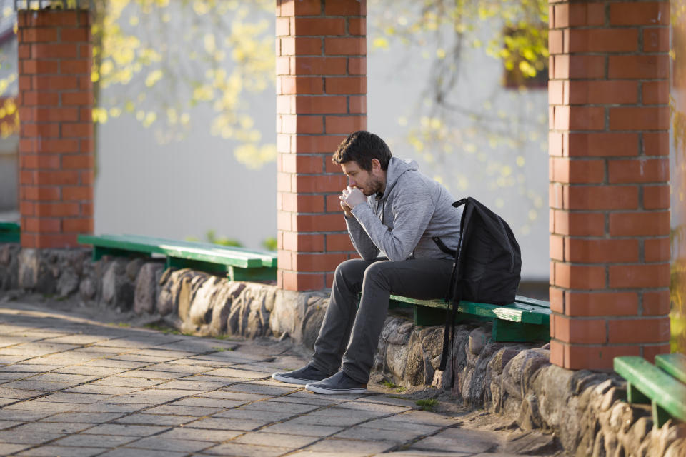 A young white man wearing a grey hoodie and jeans, with a black backpack beside him, sits on a bench with a worried expression on his face