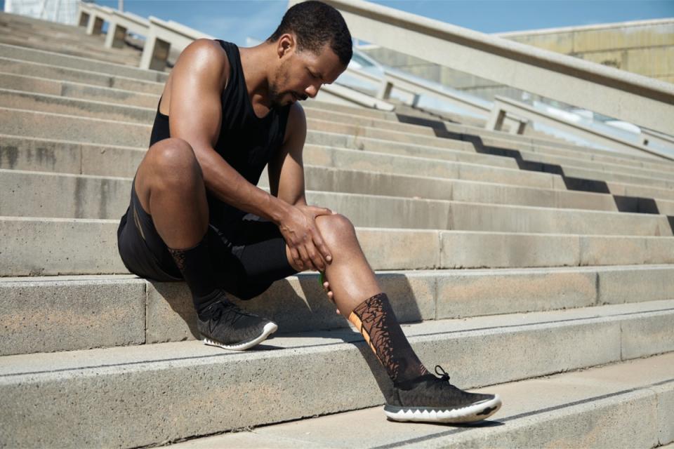 Black male jogger in black sportswear and athletic shoes sitting on stair outdoors clutching his aching knee