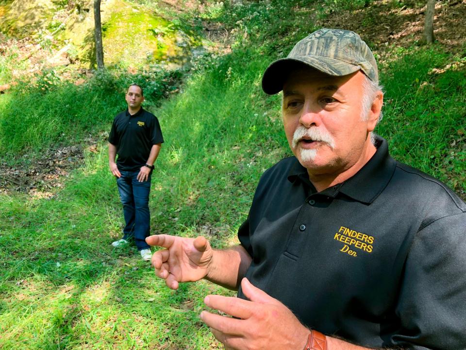 Dennis Parada stands in the foreground wearing a “finders keepers” shirt. His son stands in the background at the bottom of a grassy hill.