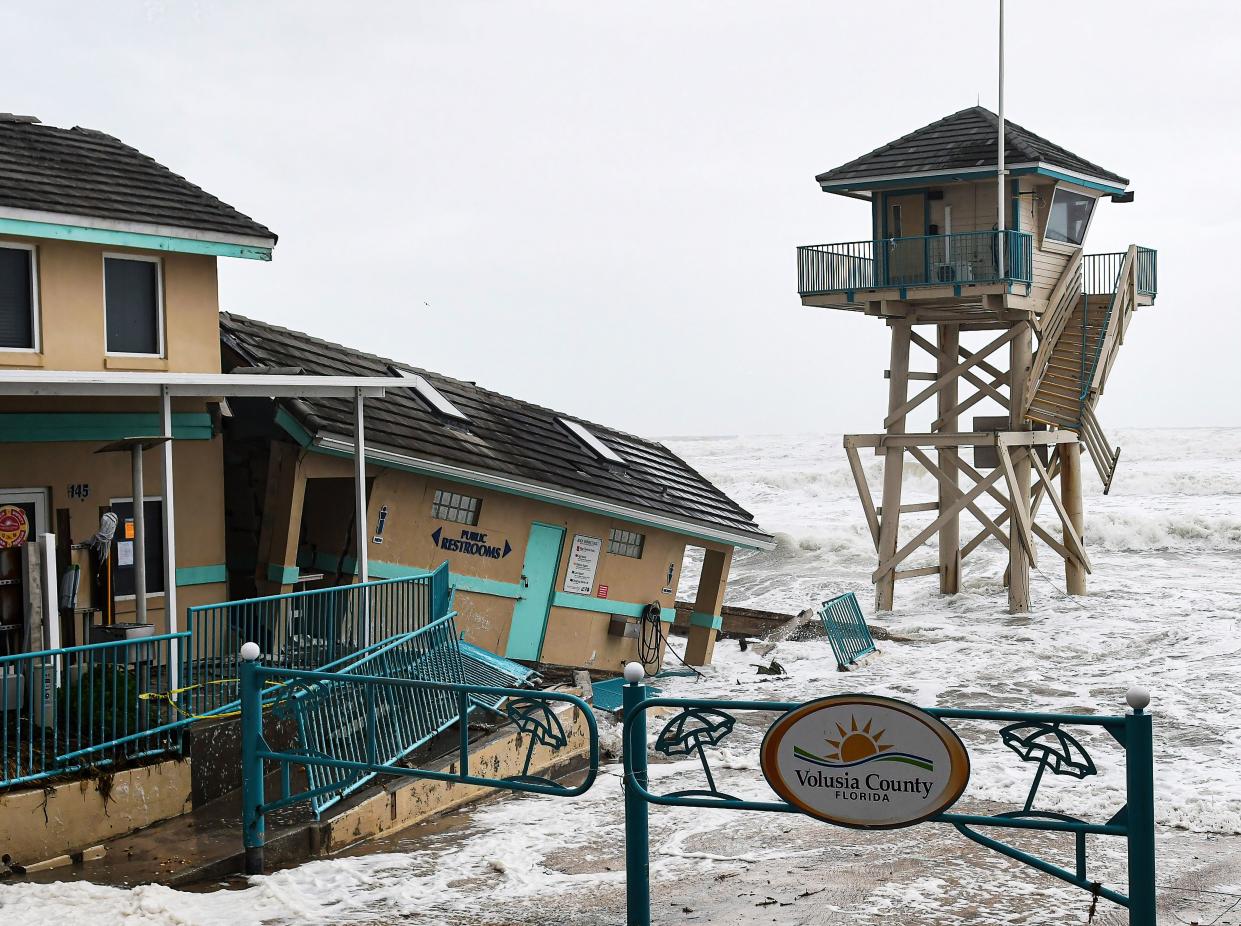 Waves crash onto a damaged building next to a lifeguard tower whose support structure is under water, with a sign surrounded by cutouts of beach umbrellas saying: Volusia County Florida.