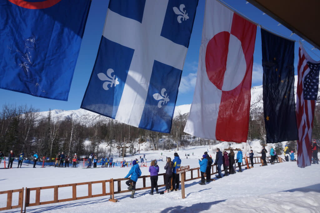 Flags of the nations with delegations to the Arctic Winter Games, along with the Alaska flag, hang from the chalet while cross-country skiers race by at Government Peak Recreation Area. The weeklong event drew 2,000 competitors from Alaska, Canada, Greenland and northern Scandinavia. (Photo by Yereth Rosen/Alaska Beacon)