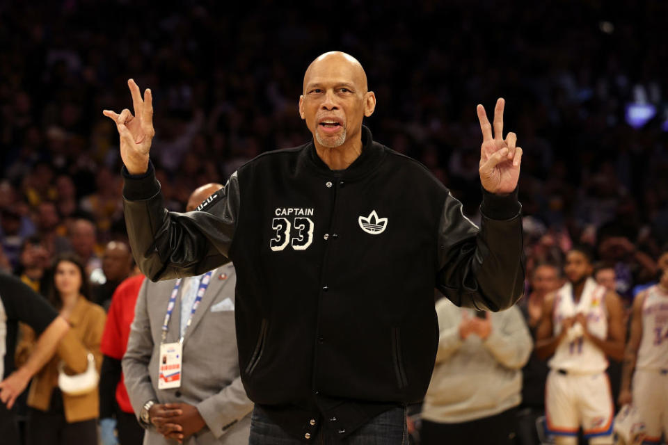 Kareem Abdul-Jabbar on a basketball court, wearing a black 'CAPTAIN 33' jacket, gesturing peace signs with both hands