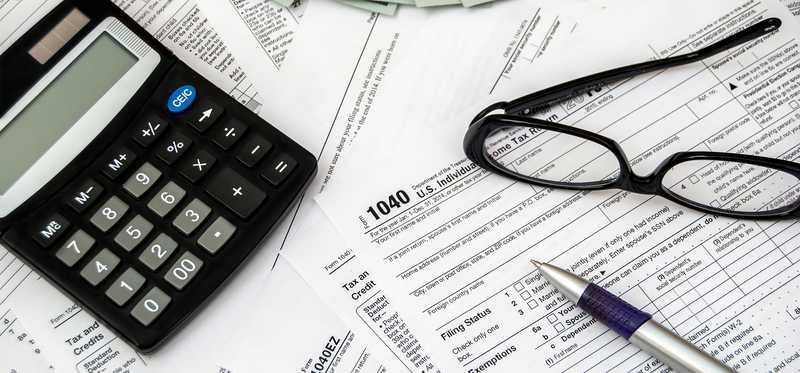 Tax forms laying scattered with a calculator, glasses, some cash, and a pen on top.