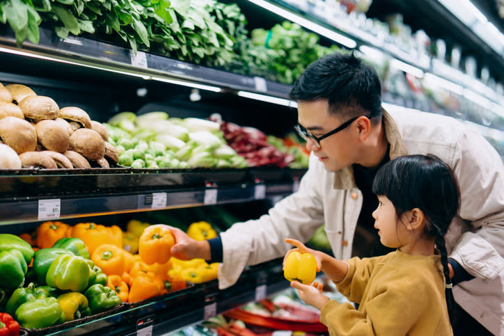 A man and a child are grocery shopping, choosing bell peppers from a supermarket vegetable section. The man is pointing out a pepper while the child holds one