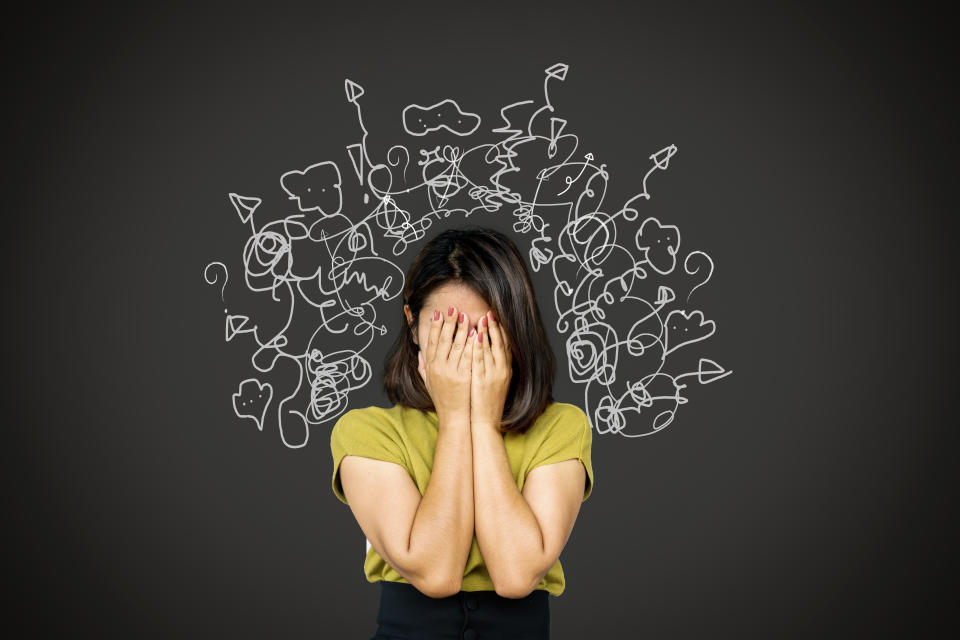 Woman with head in hands, standing in front of a blackboard covered in squiggly lines representing mental overwhelm and stress.