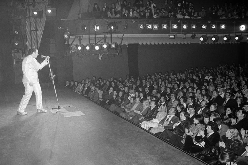 En 1975, Coluche triomphe à l’Olympia avec son deuxième spectacle, <em>Mes adieux au music-hall.</em> Sa célèbre salopette fait alors le tour de la France. Il ne s’autorise qu’une pause pour convoler en justes noces à l’automne avec Véronique. Une simple formalité pour un couple qui est déjà parent d’un petit Romain, 3 ans, et qui accueillera dès l’année suivante un petit-frère, Marius. (<em>Daniel SIMON/Gamma-Rapho via Getty Images</em>)