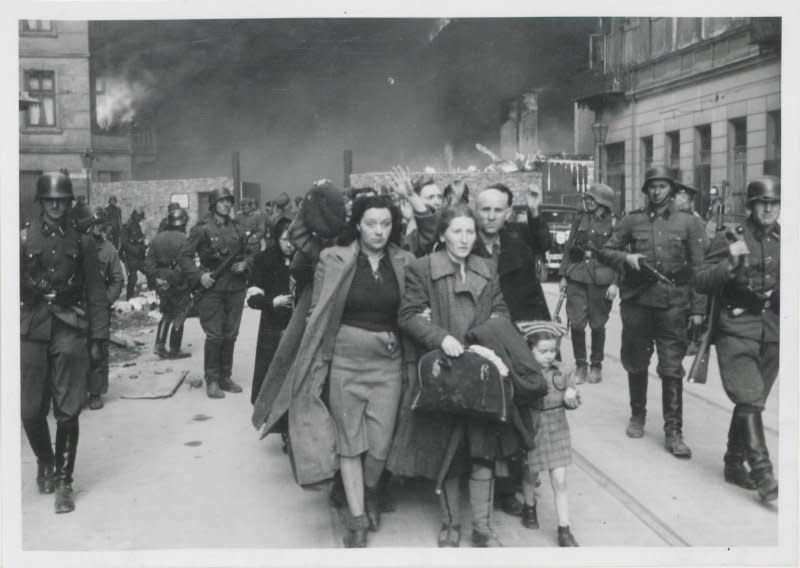 WARSAW, 1943: SS troops bring a group of captured Jewish people, including women and young children, to a railway-station collection point for deportation to the Nazi death camps; in the background, police and soldiers can be seen watching the Warsaw Ghetto burn.<span class="copyright">National Archives</span>
