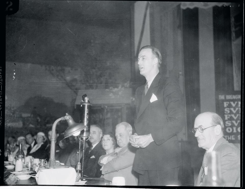 Howard Scott, man with his hair slicked back and wearing a suit stands up and addresses a crowd seated at a long wooden table.