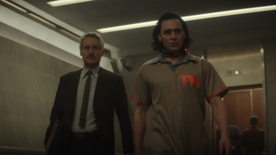 Mobius (in a suit) and Loki (in a prison uniform) walk down a hallway together.