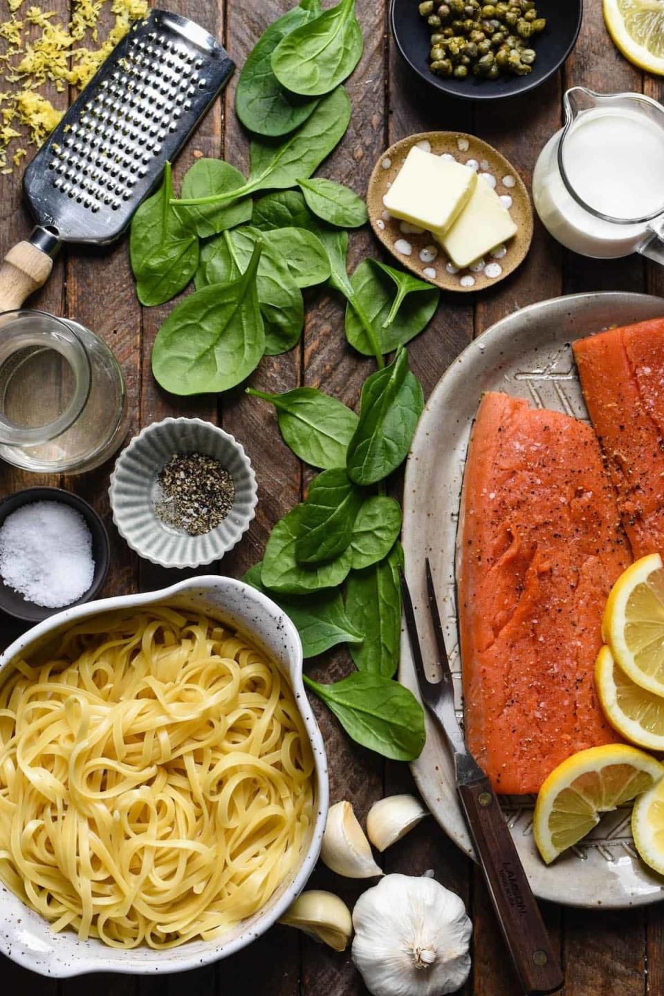 Ingredients for a pasta dish include uncooked pasta, salmon, spinach, lemon slices, garlic, capers, grated cheese, butter, and seasonings on a wooden surface