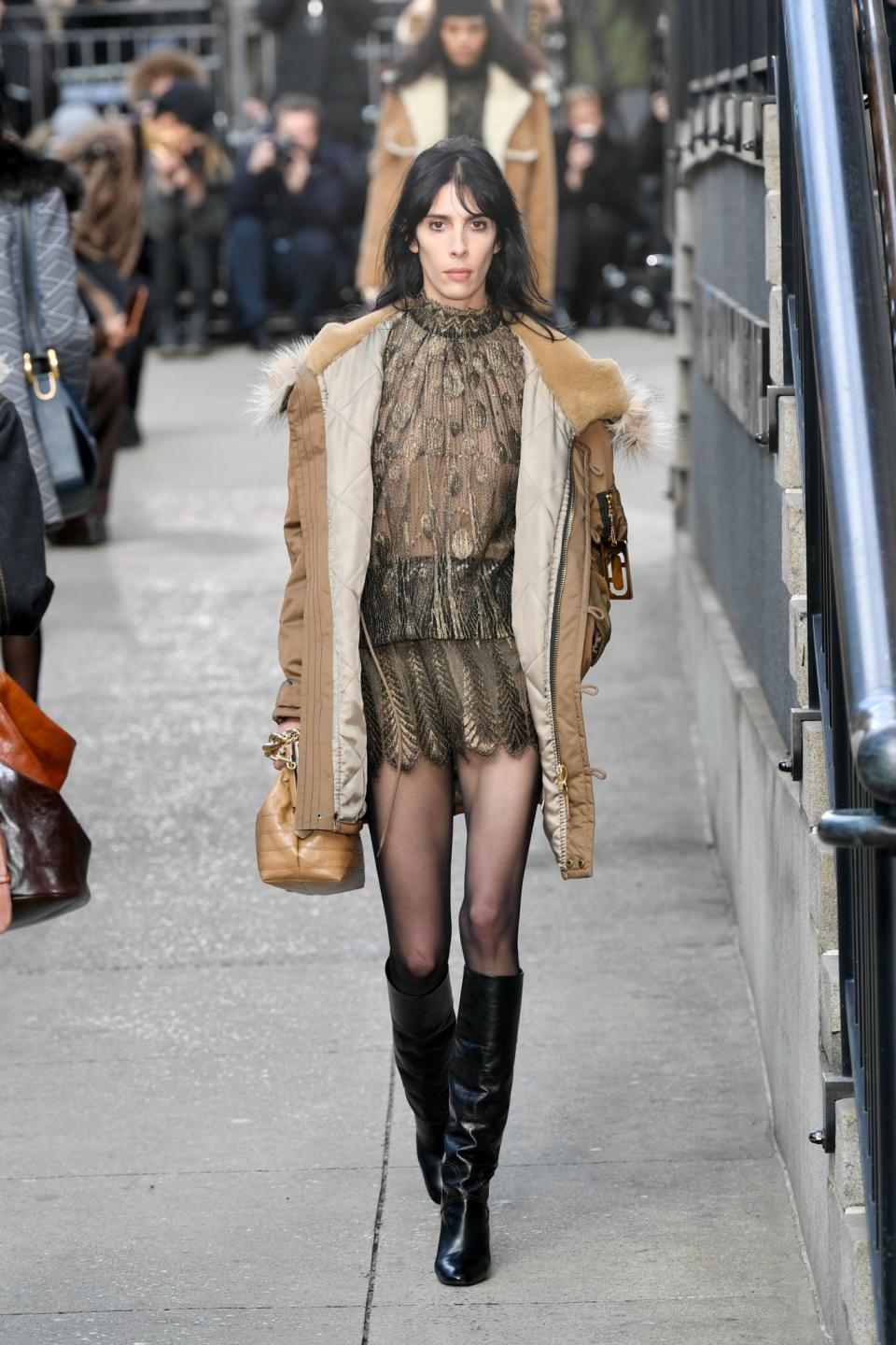 Model Jamie Bochert wears a gold embroidered semi-sheer dress and camel parka