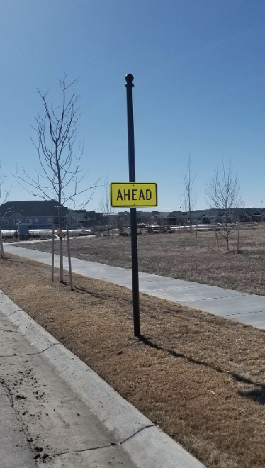 Traffic sign labeled "AHEAD" on a post beside a sidewalk with houses in the background