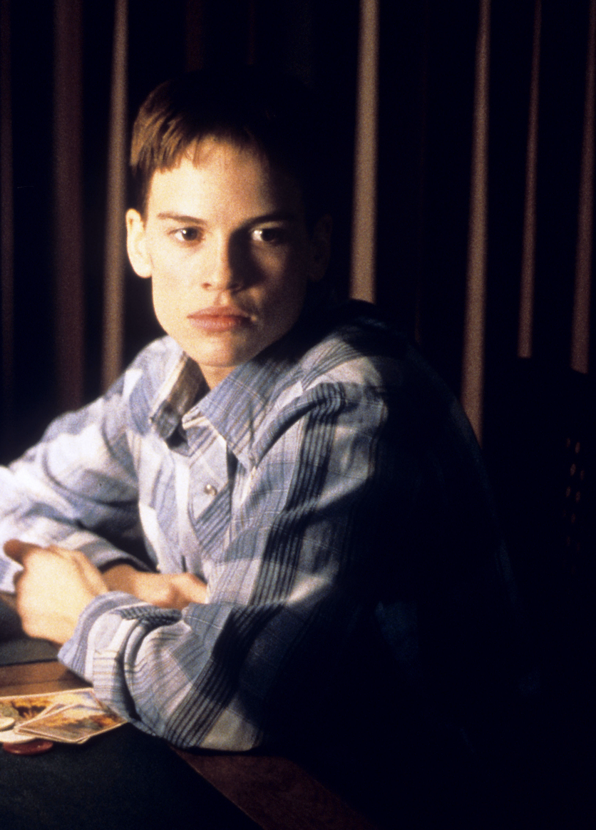 Hilary Swank portraying Brandon Teena in a plaid shirt, seated with hands clasped and looking thoughtful