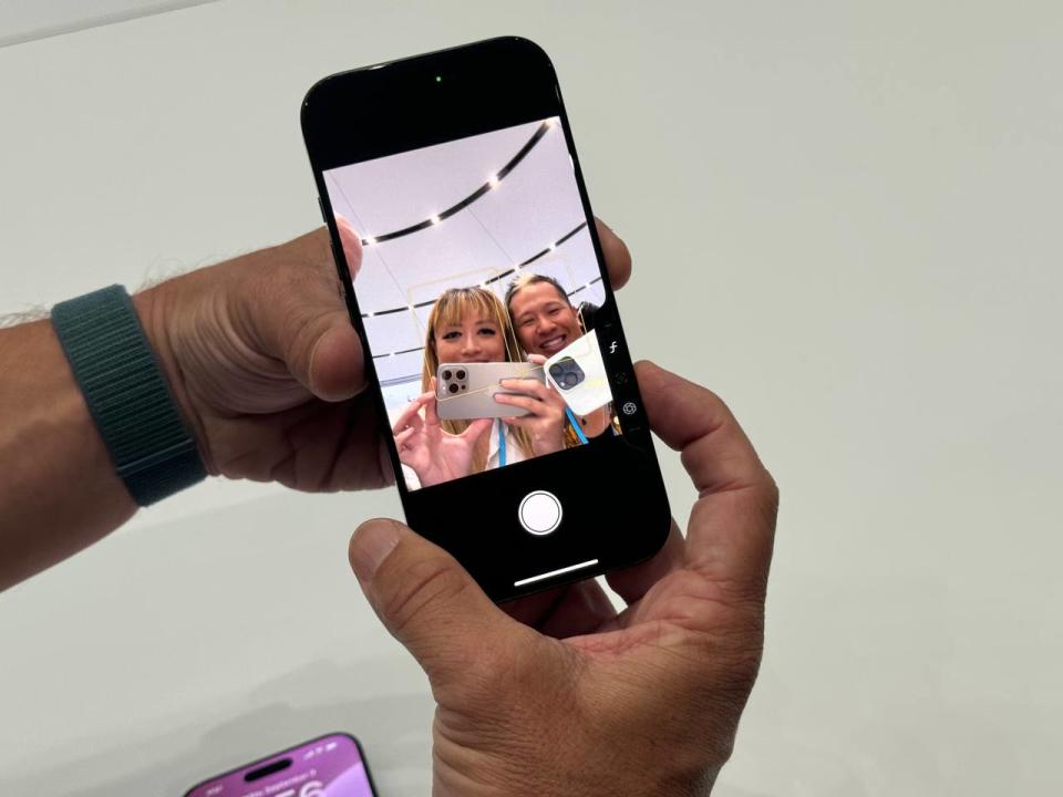 Das iPhone 16 Pro mit eingeschalteter Selfie-Kamera und zwei Personen, die auf das Telefon blicken. Der Finger einer Person liegt auf der neuen Kamera-Steuertaste unten rechts.