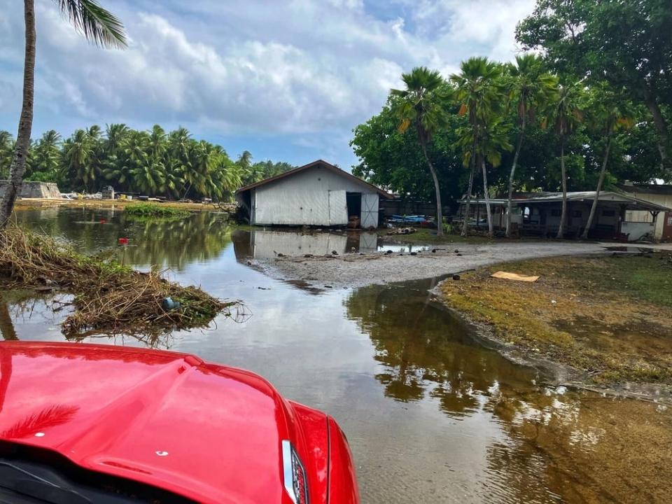 the hood of a red vehicle is visible surrounded by puddles of water with a damaged maintenance shop in the background