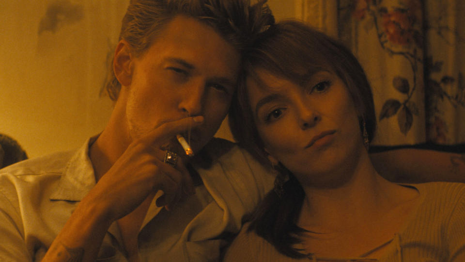 Austin Butler smokes a cigarette while sitting on a couch next to Jodie Comer in The Bikeriders.