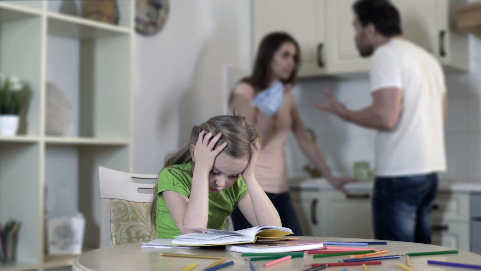 Child looks stressed studying while adults argue in the background, depicting family tension affecting children's well-being