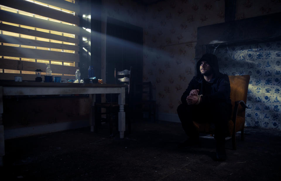 Aaron Dingle in a hood as he sits in a dark abandoned room on a chair.