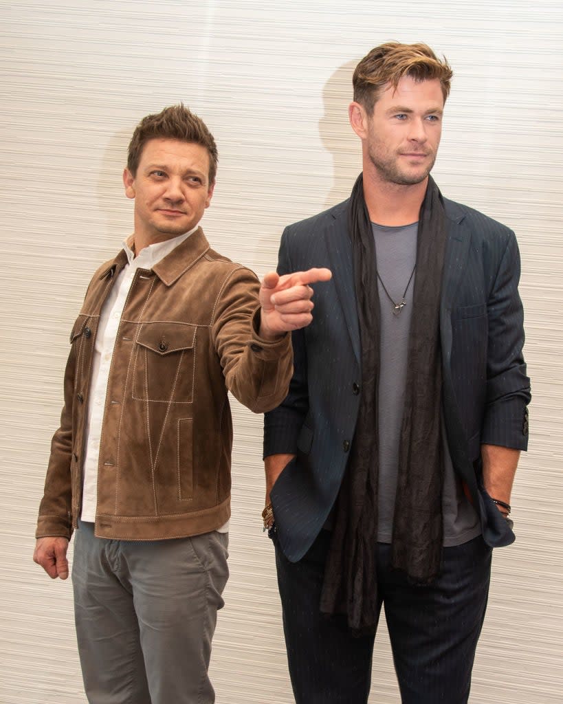 Jeremy Renner is pointing while standing next to Chris Hemsworth, who is wearing a blazer and scarf