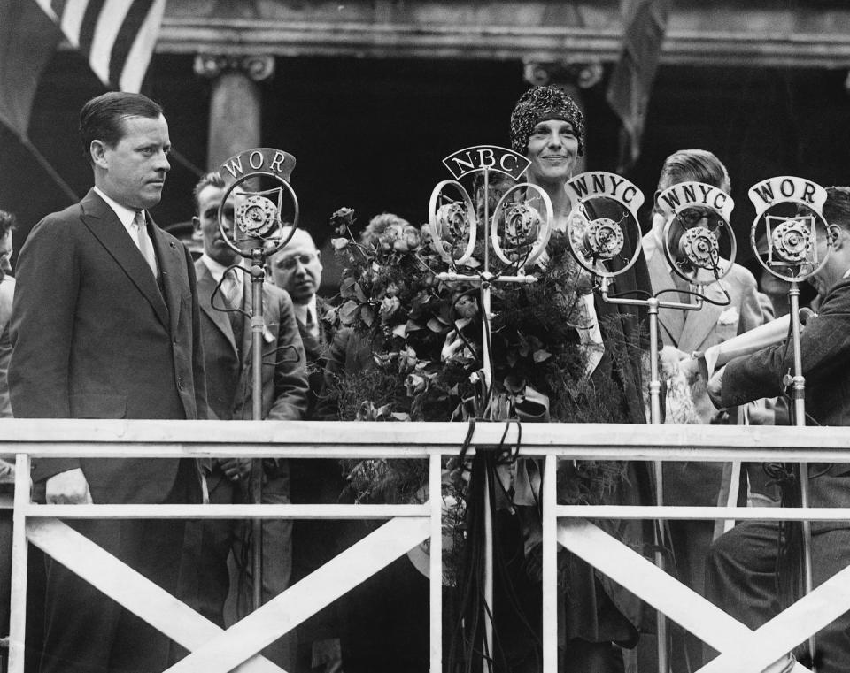 amelia earhart smiles and stands behind several microphones, she holds a large bouquet and wears a cap and dress