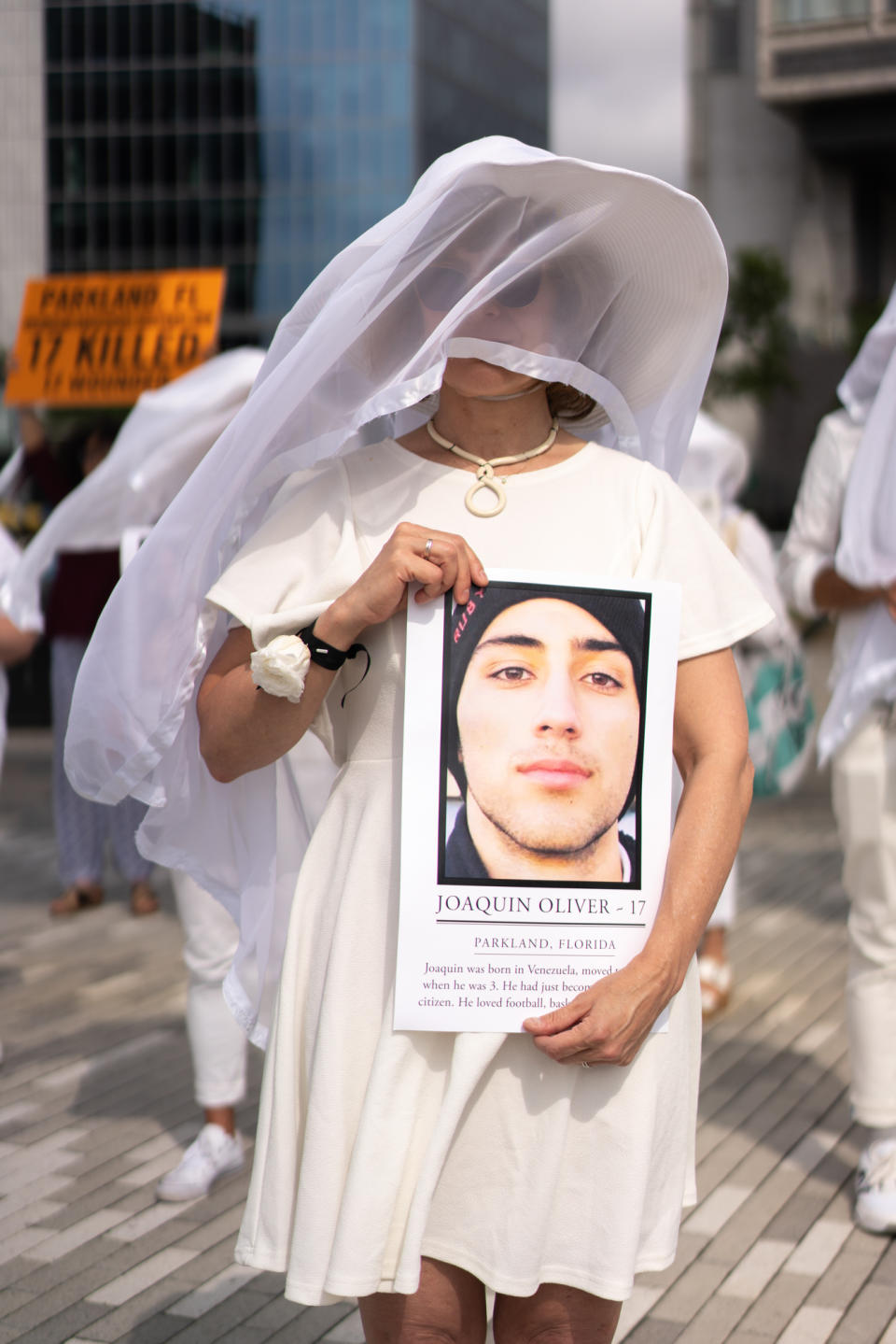 A person wearing white with a veil holds a picture of Joaquin Oliver with writing below that reads: Joaquin Oliver, 17, Parkland, Florida. (Followed by information about Joaquin's life.)