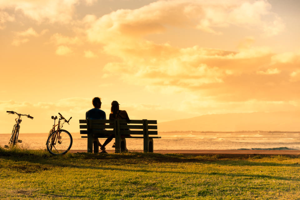 A couple sits on a bench by the ocean with two bicycles nearby, enjoying a serene sunset