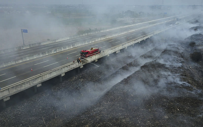 From a distance, looking slightly down, is seen a highway that extends from the upper right side of the image to the lower left. The ground on either side of the highway is dark brown, with lots of smoke rising from the charred ground. In the center of the image, parked on the side of the highway, is a red fire engine. Beside it, a firefighter is spraying a stream of water from the highway toward the ground below