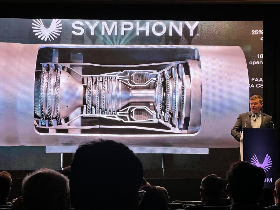 Boom Supersonic's Symphony engine on a screen display, in front of which stands CEO Blake Scholl at a company podium.