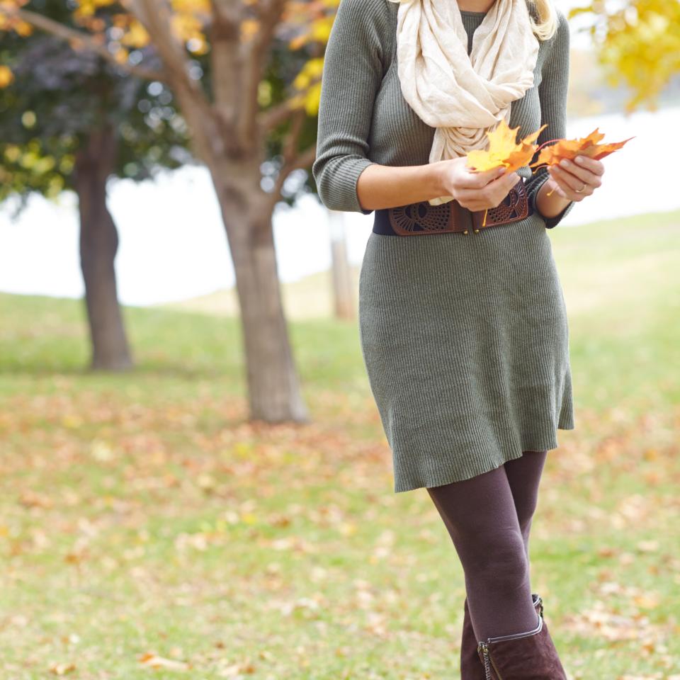 A woman stands outdoors in a park, wearing a knit dress, leggings, knee-high boots, a scarf, and a flat cap, holding autumn leaves, smiling and looking down