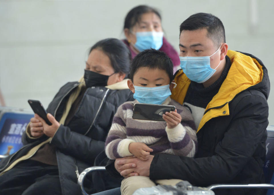 A father and son wear masks in Hong Kong airport as flights are delayed. The little boy is sitting on his father's lap holding a phone.