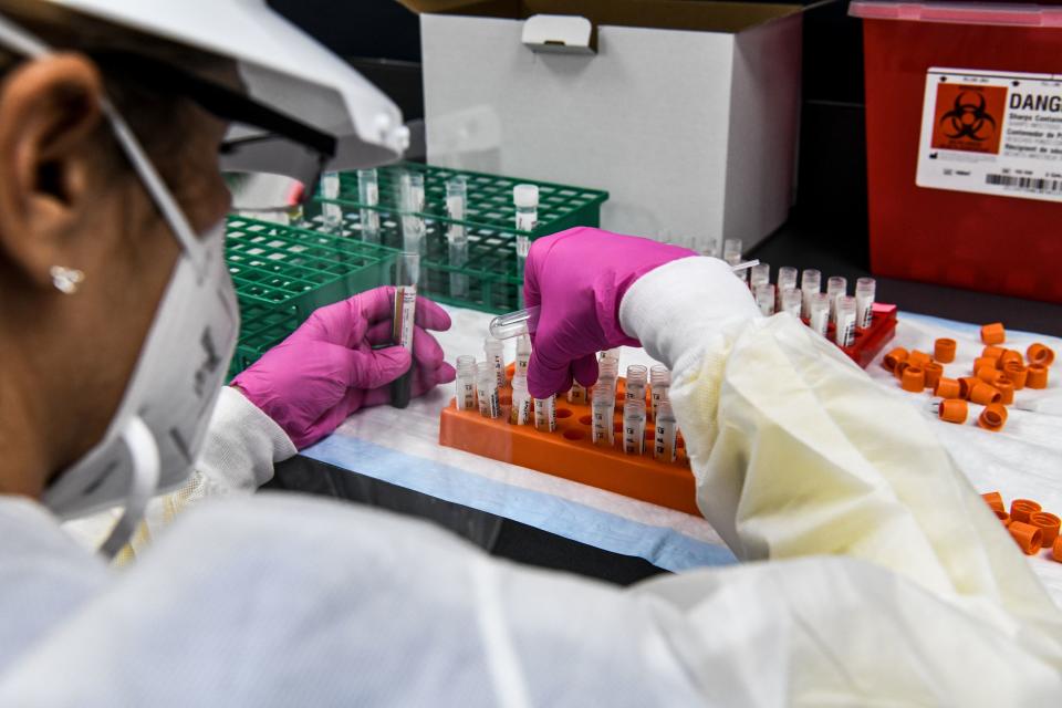 A lab technician wearing a face mask and pink rubber gloves holds glass vials