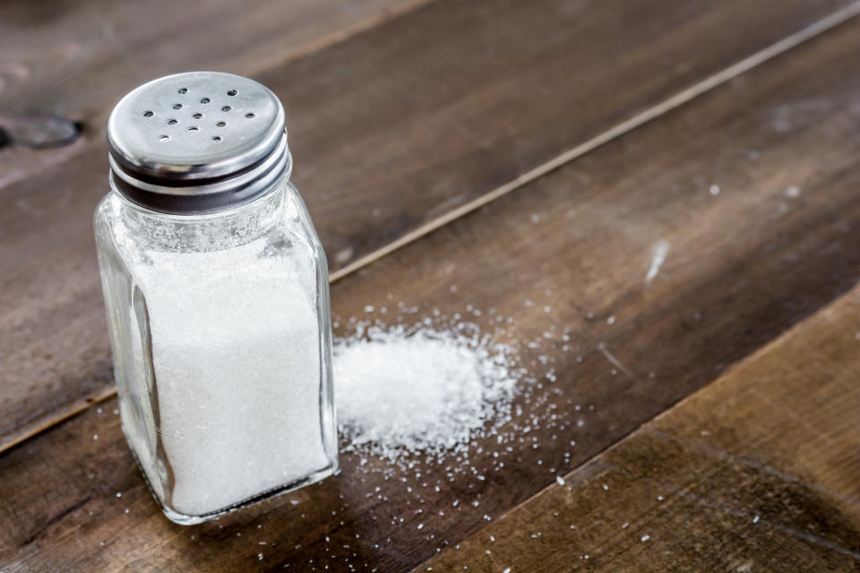 Salt shaker sitting on a wood table with a loose pile of salt on the side.