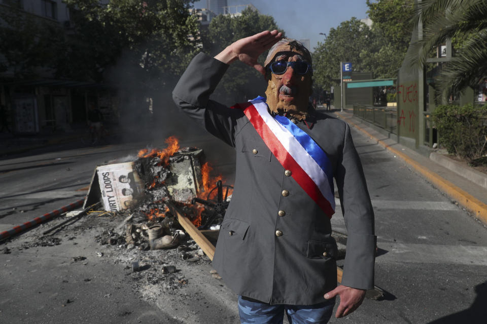 FILE - In this Nov. 6, 2019 file photo, an anti-government demonstrator costumed as late military dictator Gen. Augusto Pinochet salutes in front of a burning street barricade placed by protesters demanding a new constitution in Santiago, Chile. Chileans will elect an assembly on April 11, 2021, tasked with writing fresh governing principles and putting them to a national vote in the first half of 2022 to replace the much-amended relic of military rule, the 1980 constitution. (AP Photo/Esteban Felix, File)