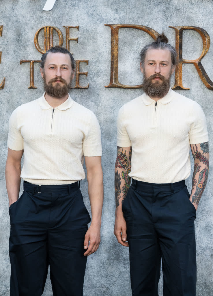 Ryan and Adam Metzger standing side by side, wearing matching white shirts and dark pants. Adam has tattooed arms, Ryan does not. Both have beards and tied-back hair