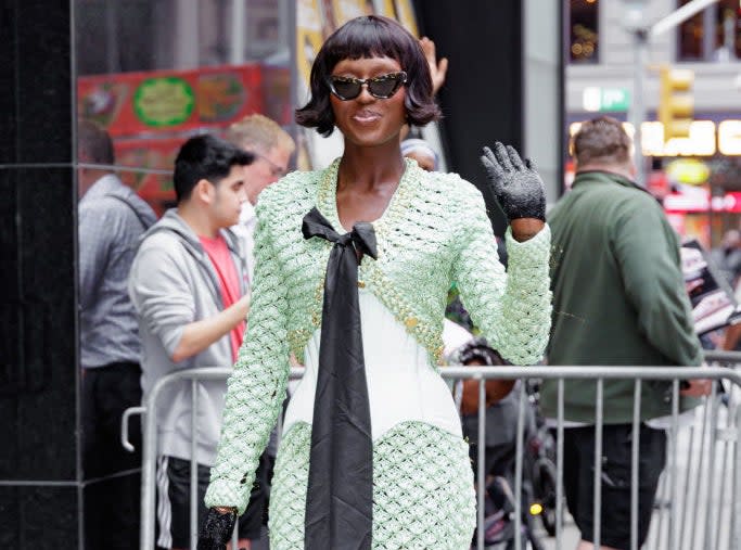 Jodie in a textured gown with a long bow accessory waves to the crowd on the sidewalk