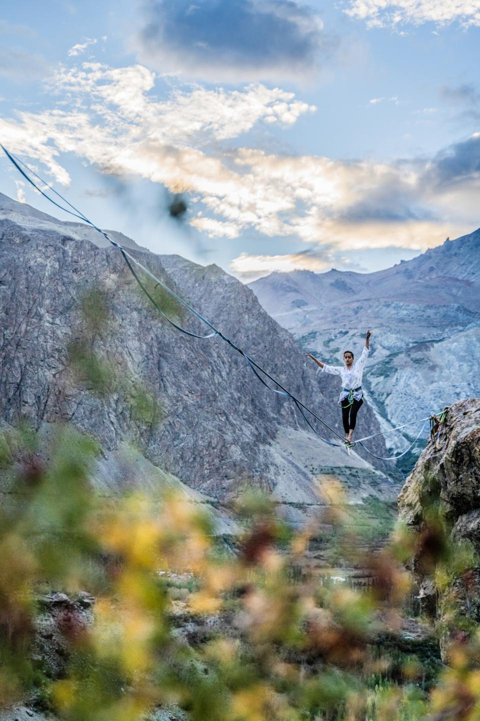 A woman with arms up and outstretched balances on a slack line over a rocky ravine with mountains in the background.