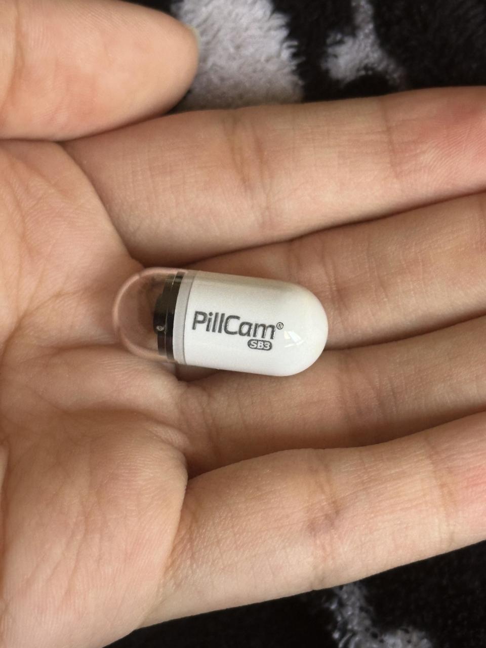 A hand holds a small capsule labeled "PillCam SB3," typically used for medical imaging