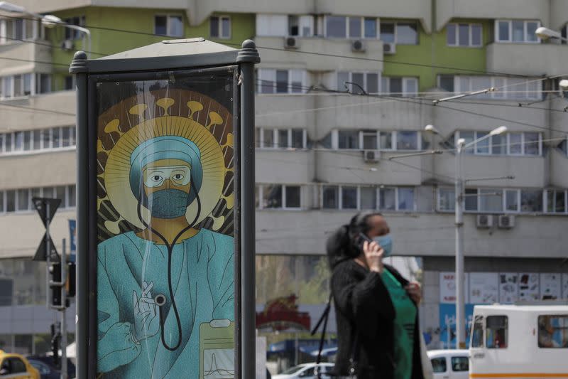 A woman wearing a medical mask passes by an outdoor poster that shows a medic depicted in a similar manner as Jesus Christ would be portrayed by Christian Orthodox religious paintings