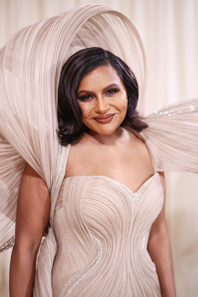 Mindy Kaling at a formal event, wearing an intricately designed gown with structured, flowing details around her shoulders