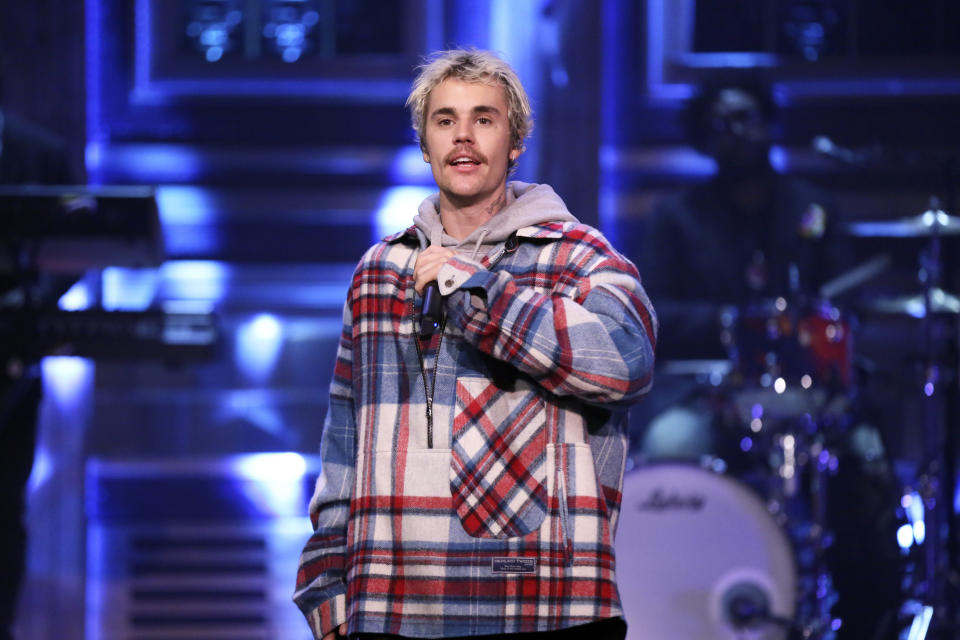 Justin Bieber performing on stage, wearing a plaid jacket over a hoodie. Musicians and instruments are visible in the background