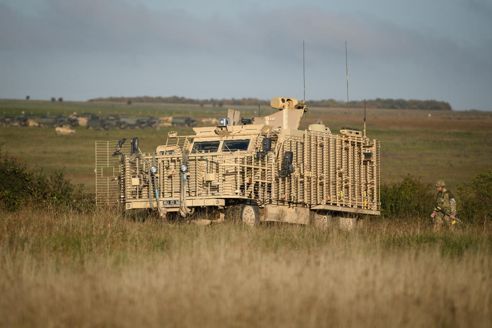 A soldier approaches the rear of a Mastiff armored vehicle in a grassy field with other vehicles in the far distance.