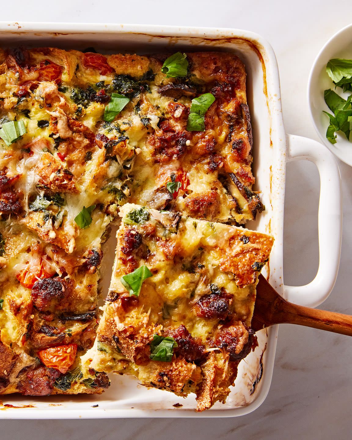 eggy breakfast strata featuring sausage, tomatoes, bread, and mushrooms, and topped with basil