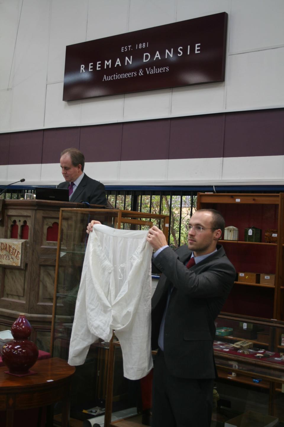 An auctioneer holds up a pair of linen drawers that belonged to Queen Victoria, with a blue embroidered cipher and a 38" waist.  The bloomers were won by a collector in Essex (Reeman Dansie)